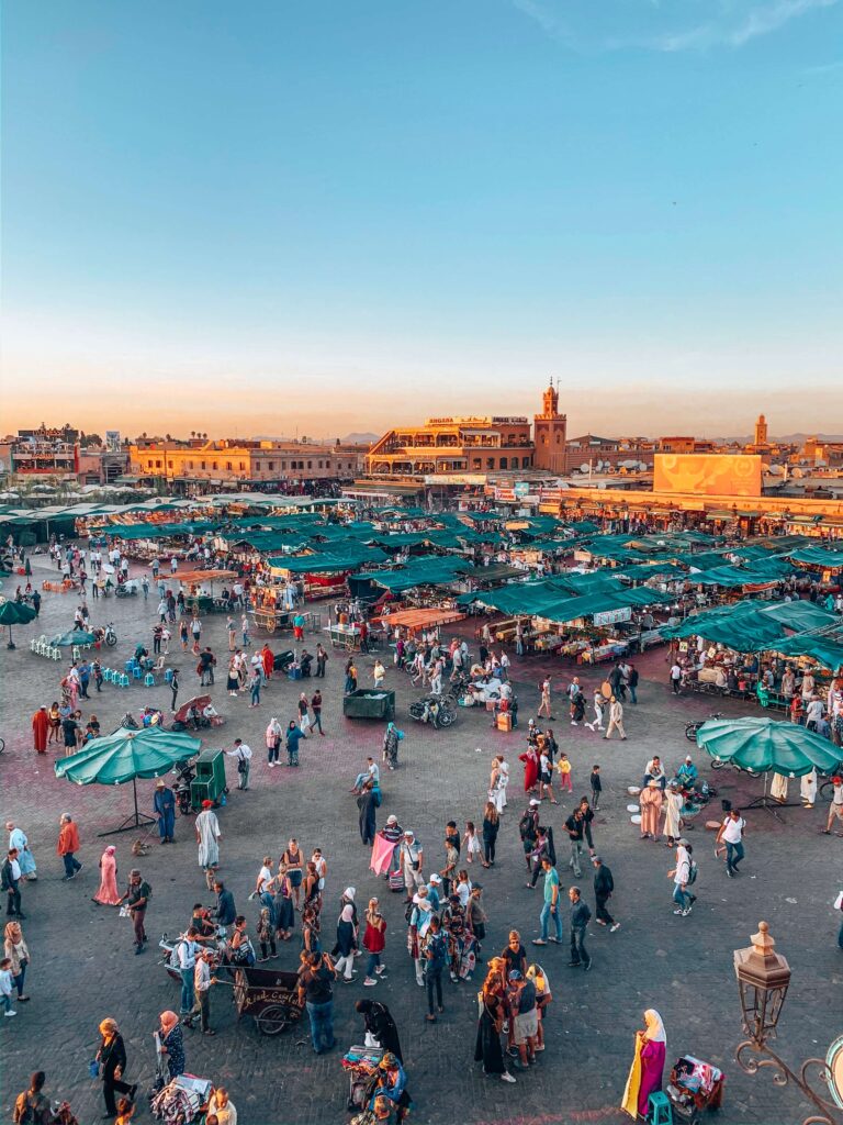 12 Charming Budget Riads in Marrakech 3 A bustling outdoor market square filled with people, green vendor tents, and stalls at sunset. The sky is clear, historic buildings line the background—reminiscent of evenings near budget riads in Marrakech.