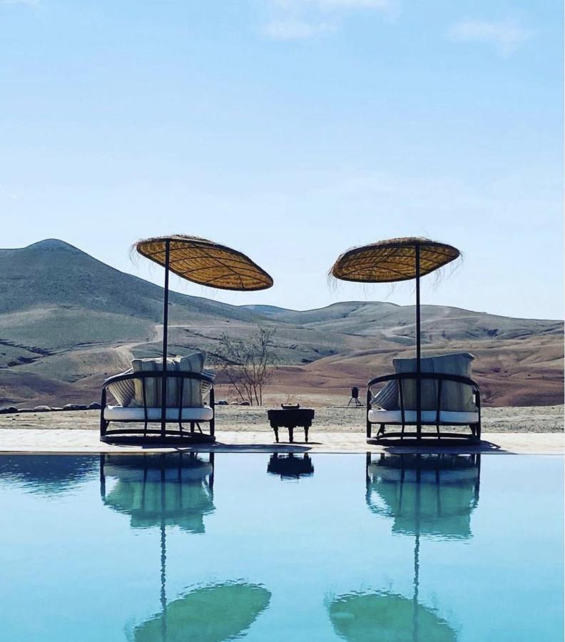 8 SPECTACULAR Agafay Desert Camps 13 Two lounge chairs with straw umbrellas are reflected in a still swimming pool, set against arid hills and a clear blue sky—evoking the tranquil and inviting atmosphere of Agafay Desert camps.