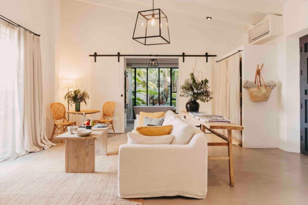 Unique Agroturismos in Mallorca 31 Bright, cozy living room inspired by agroturismos in Mallorca, featuring a white sofa, wooden coffee and dining tables, wicker chairs, and large windows with sheer curtains. Green plants and natural light create a warm, inviting atmosphere.