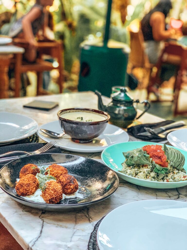 10 Unique Riads in Marrakech with Pool 46 A marble table set with various dishes, including fried balls on sauce, a bowl of soup, and a salad topped with avocado and tomato—reminiscent of meals enjoyed at budget riads in Marrakech; people dine in the blurred background.