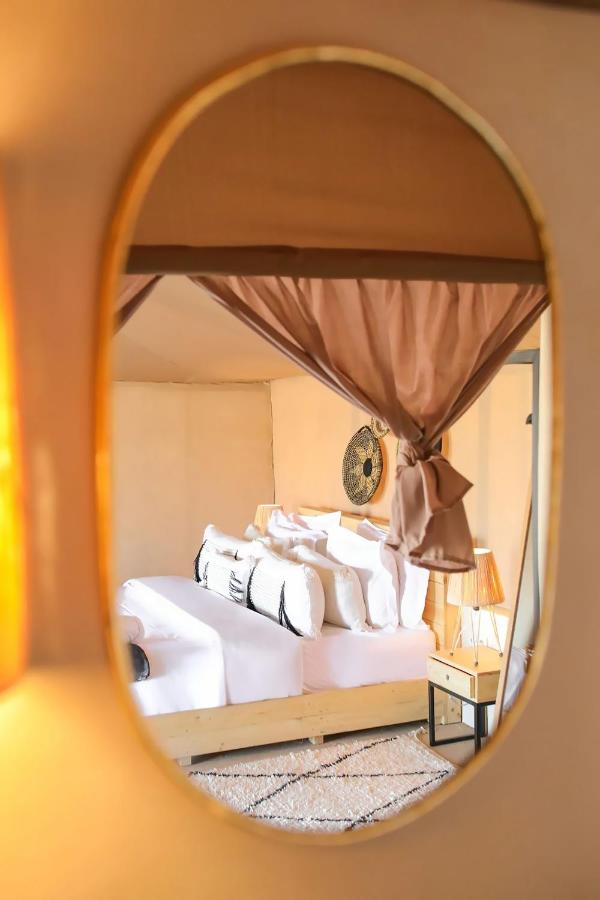 8 SPECTACULAR Agafay Desert Camps 42 Reflection in an oval mirror reveals a cozy bedroom reminiscent of agafay desert camps, with a neatly made bed, white linens, decorative pillows, a wooden headboard, bedside tables, and warm ambient lighting.