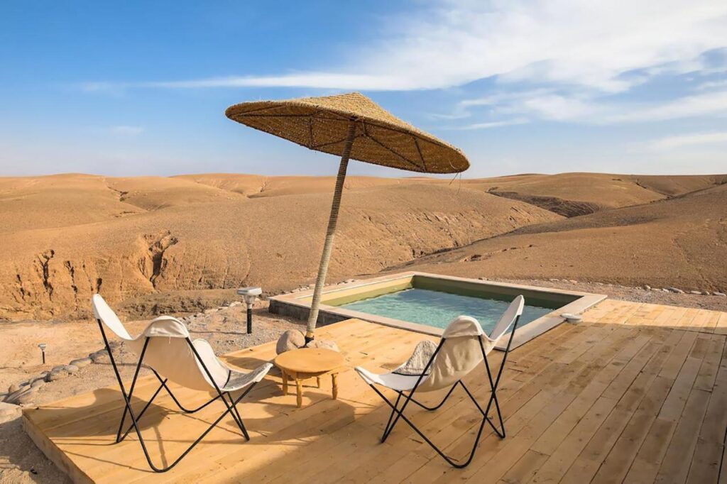 8 SPECTACULAR Agafay Desert Camps 40 Two white lounge chairs and a small table sit under a large straw umbrella on a wooden deck beside a small pool, overlooking the vast Agafay Desert camps and dry landscape under a clear blue sky.