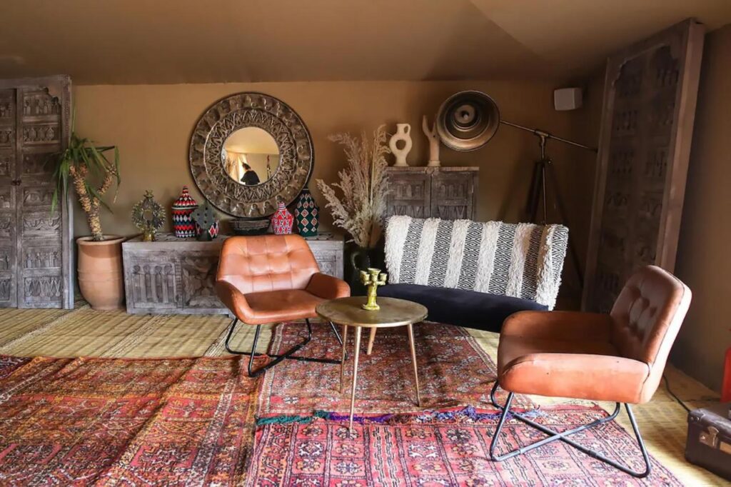 8 SPECTACULAR Agafay Desert Camps 41 A cozy living room with brown leather chairs, a round coffee table, patterned rugs inspired by Agafay Desert camps, a decorative mirror, rustic cabinets, potted plants, and various decorative items on display.