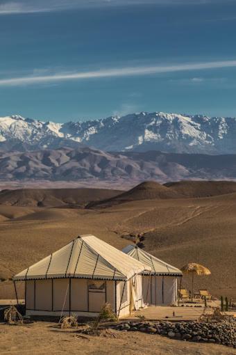 8 SPECTACULAR Agafay Desert Camps 39 A white tent stands in a dry, brown Agafay Desert Camps landscape with snow-capped mountains in the distance under a blue sky.