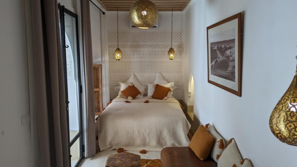 12 Charming Budget Riads in Marrakech 42 A cozy bedroom with a double bed, white and beige bedding, brown accent pillows, hanging lanterns, a small sofa, and framed art on the wall, reminiscent of warm Moroccan-inspired decor found in many budget riads in Marrakech.