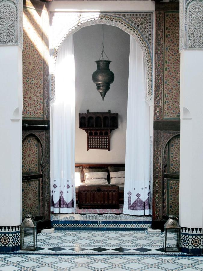 7 UNIQUE Riads in Fez You Won't Forget 4 A Moroccan-style interior reminiscent of riads in Fez, with intricate mosaic tiles, arched doorway, white curtains, a hanging metal lantern, and traditional wooden furniture, including a cushioned bench and carved wall shelf.