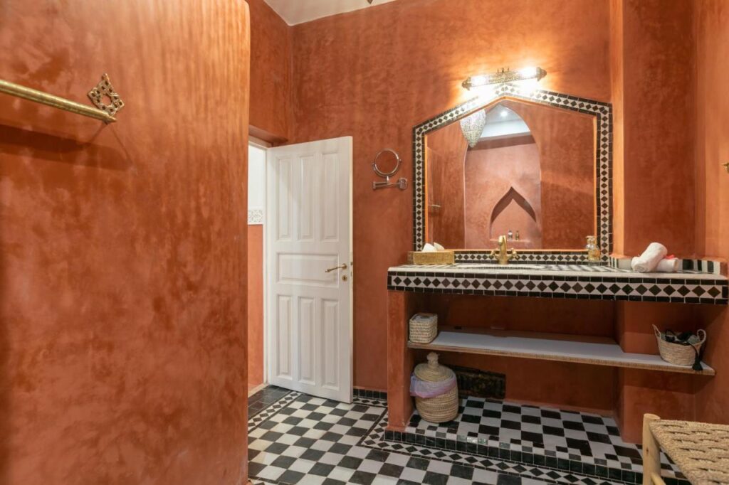 12 Charming Budget Riads in Marrakech 40 A bathroom inspired by budget riads in Marrakech, with warm terracotta walls, a white door, and a black-and-white checkered floor. The vanity has a large mirror framed by geometric tiles and woven baskets on open shelves below the sink.