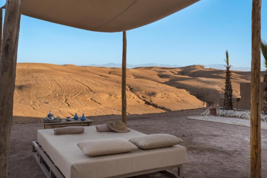 8 SPECTACULAR Agafay Desert Camps 17 A shaded outdoor bed with cushions sits on sandy ground, overlooking the vast Agafay Desert camps under a clear blue sky. A small table with tea sets and a straw hat rest nearby, while sparse palm trees dot the distant horizon.