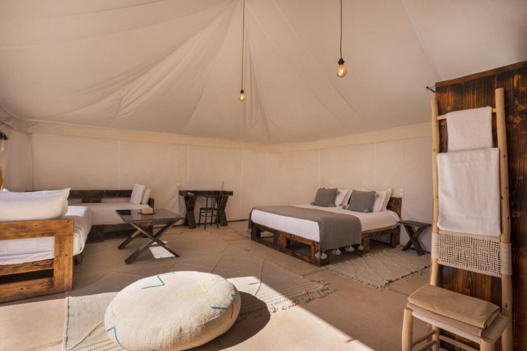 8 SPECTACULAR Agafay Desert Camps 16 A spacious glamping tent interior, inspired by Agafay desert camps, features wood-framed beds, neutral bedding, a small desk, cushioned pouf, towel rack, soft lighting, and woven rugs. The decor is simple and cozy.