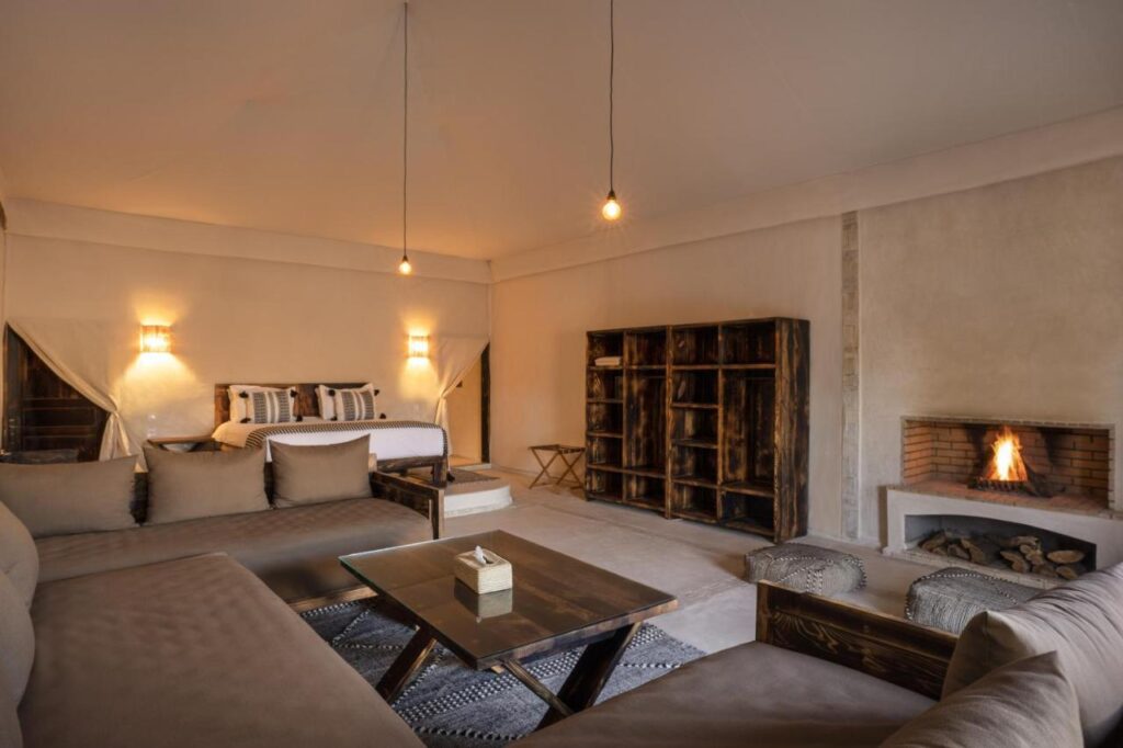 8 SPECTACULAR Agafay Desert Camps 18 A cozy, rustic bedroom and living area reminiscent of agafay desert camps, with earth-toned walls, a bed with striped pillows, a sectional sofa, wooden shelves, a coffee table, and a lit fireplace softly illuminated by hanging light bulbs.