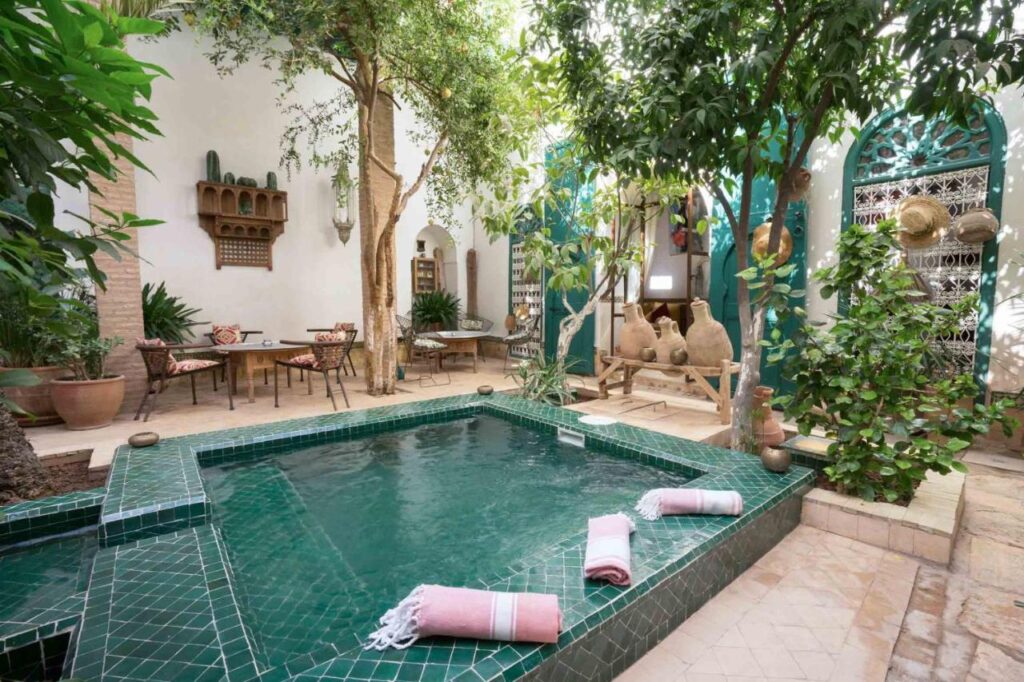 12 Charming Budget Riads in Marrakech 10 A serene courtyard with a green-tiled plunge pool, surrounded by lush plants and traditional decor—typical of budget riads in Marrakech. Rolled towels by the pool and pottery accents enhance the relaxing, Moroccan-inspired ambiance.