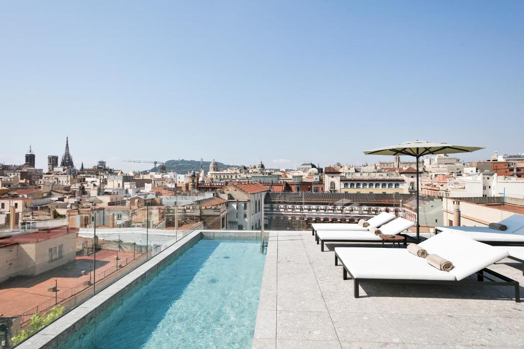 Top Barcelona Hotels with Rooftop Pool 12 Rooftop pool with lounge chairs and towels overlooking a cityscape of historic buildings and church spires under a clear blue sky—a perfect example of the luxury found in Barcelona Hotels with Rooftop Pool.