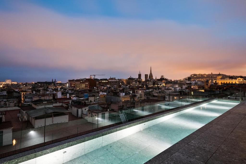 Top Barcelona Hotels with Rooftop Pool 11 A rooftop infinity pool overlooks a cityscape at dusk, with buildings, church spires, and distant hills under a colorful sky—an iconic scene reminiscent of Barcelona hotels with rooftop pools.