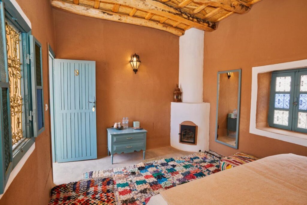 8 SPECTACULAR Agafay Desert Camps 26 A cozy room with warm brown walls evokes the charm of agafay desert camps, featuring a wooden ceiling, colorful rug, blue door, side table with drinks, wall lamp, white fireplace, mirror, and blue-framed windows with patterned glass.