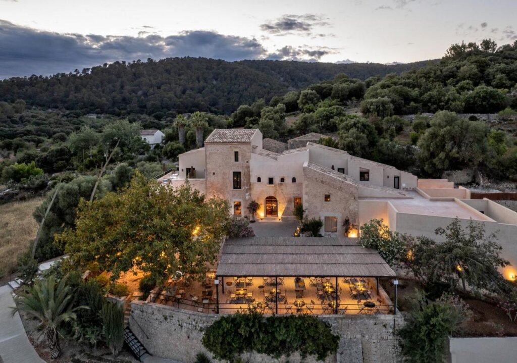 Unique Agroturismos in Mallorca 9 A rustic, stone building surrounded by trees and hills at dusk, warmly lit by outdoor lights. This inviting scene captures the charm of agroturismos in Mallorca, with a covered terrace offering cozy tables and chairs in the foreground.
