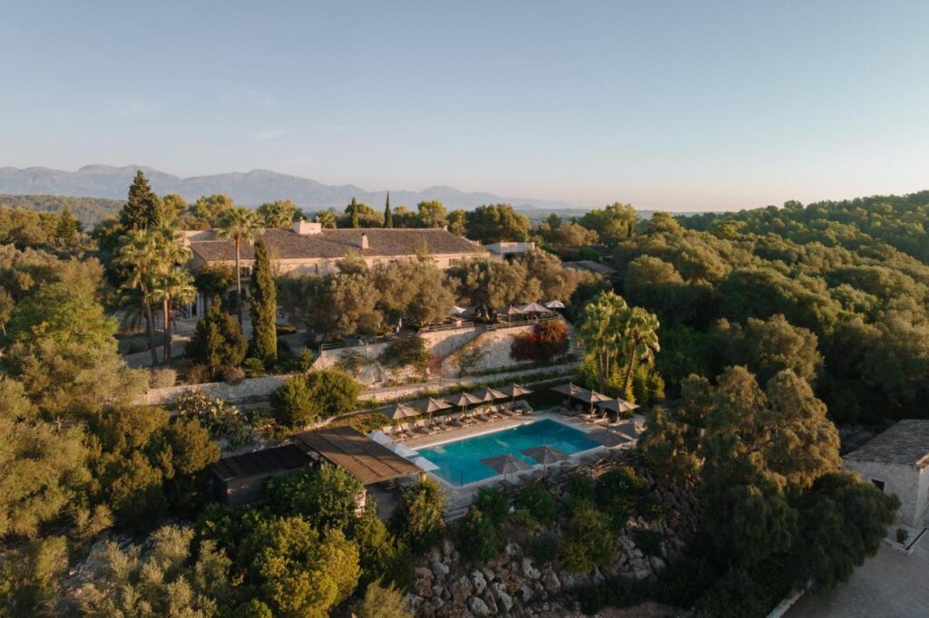 Unique Agroturismos in Mallorca 30 A large stone villa surrounded by lush greenery and trees, with a rectangular swimming pool and sun loungers in the foreground—perfect for those seeking authentic agroturismos in Mallorca, all set against distant mountain views.