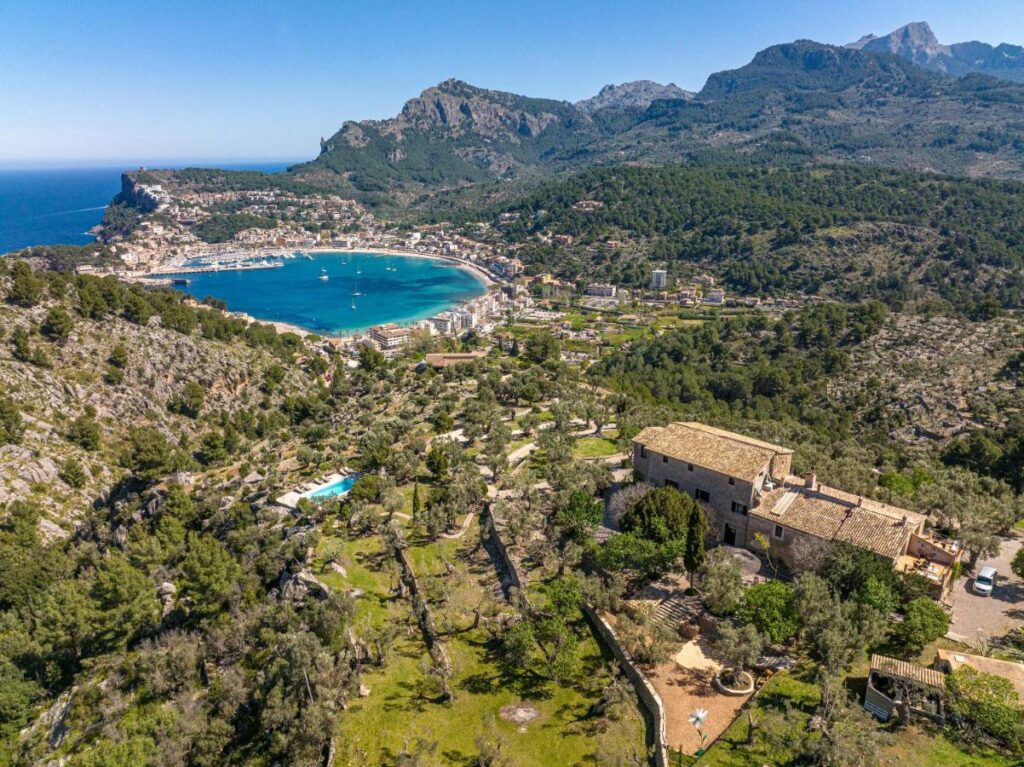 Unique Agroturismos in Mallorca 39 Aerial view of a coastal town in Mallorca with a bay, green mountains, and forests. Scattered buildings and agroturismos in Mallorca are visible, with a large stone house with a tile roof in the foreground under a clear blue sky.