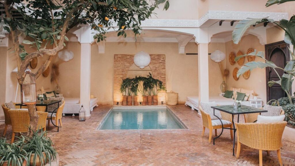 12 Charming Budget Riads in Marrakech 25 A cozy courtyard at one of the budget riads in Marrakech, featuring a small rectangular pool, wicker chairs, lush plants, and cushioned benches under a covered patio. Warm lighting and earth-toned walls create a relaxing, inviting atmosphere.