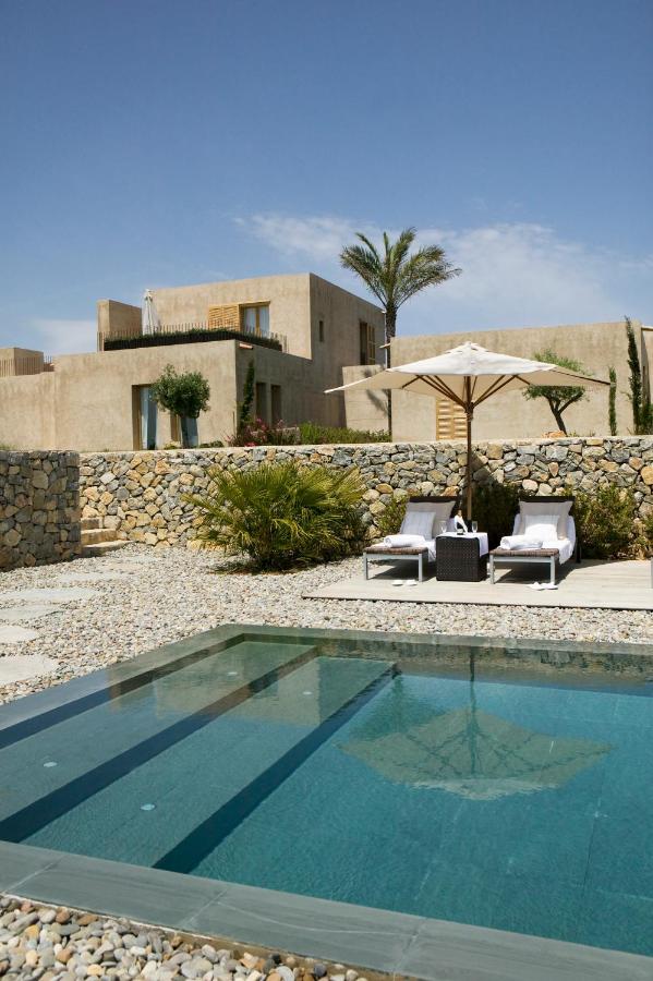 Unique Agroturismos in Mallorca 2 A modern villa with stone walls, palm trees, and two lounge chairs under a white umbrella beside a clear outdoor swimming pool on a sunny day, capturing the relaxed charm of agroturismos in Mallorca.