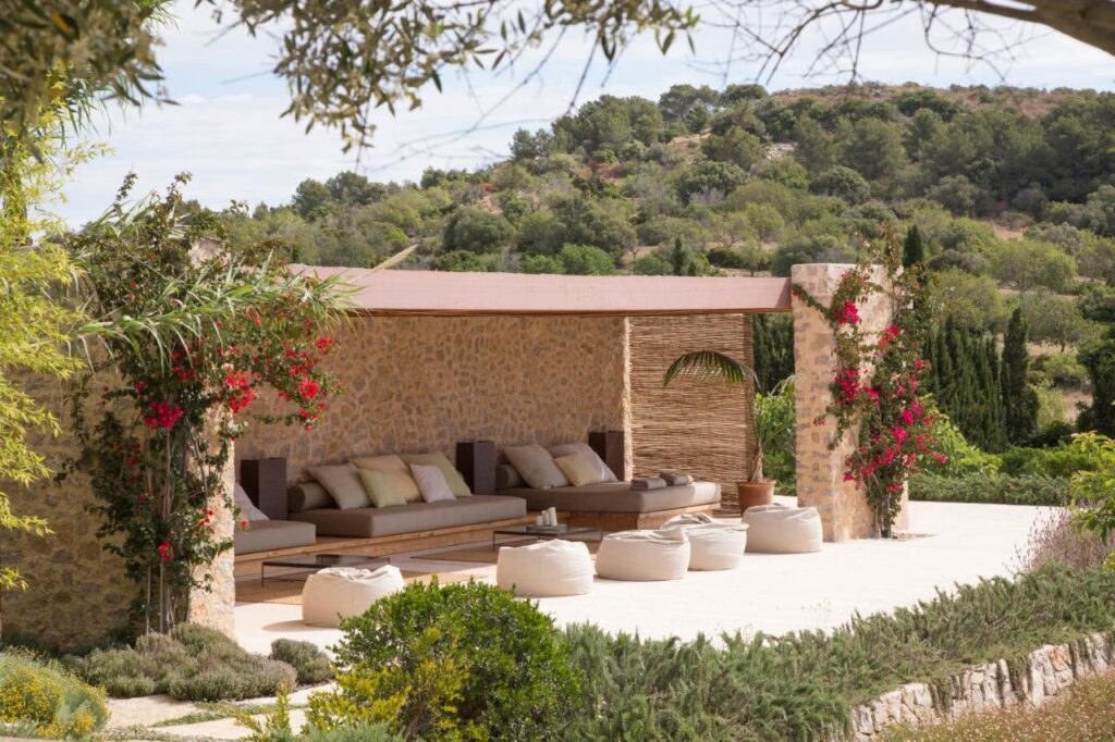 Unique Agroturismos in Mallorca 5 A shaded outdoor lounge with stone walls, plush sofas, and white bean bags is surrounded by lush greenery and vibrant climbing flowers, offering a tranquil retreat typical of agroturismos in Mallorca amid rolling hills and trees.