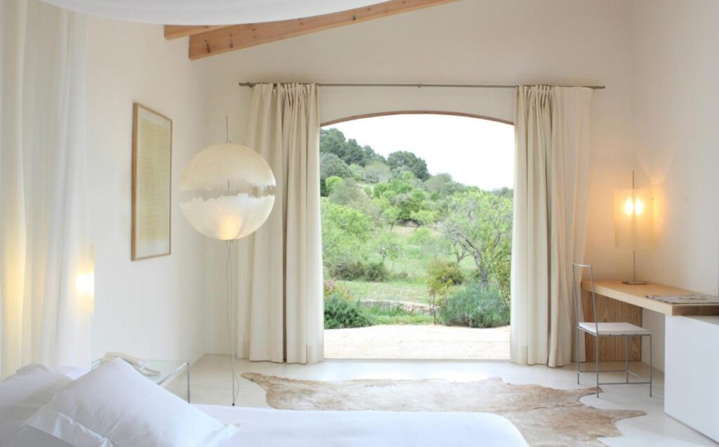 Unique Agroturismos in Mallorca 4 Minimalist bedroom with white walls, a bed, a modern globe lamp, desk and chair, and cream curtains framing a large open doorway to a lush garden view—echoing the serene style of agroturismos in Mallorca. Natural light fills the tranquil space.