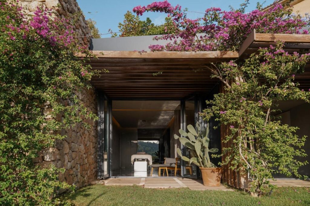 Unique Agroturismos in Mallorca 35 A modern house with stone walls and a wooden pergola, like those found in agroturismos in Mallorca, is adorned with blooming purple flowers and green vines. Sliding glass doors open to a patio with a potted cactus, a chair, and a view of the outdoors.