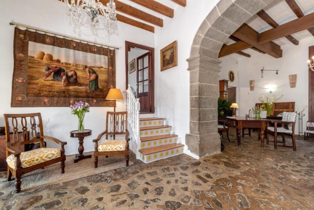 Unique Agroturismos in Mallorca 41 A rustic living room inspired by agroturismos in Mallorca, with stone floors, wooden ceiling beams, a large stone arch, patterned cushions on wooden chairs, a tapestry on the wall, tiled staircase, and a wooden dining table in the background.