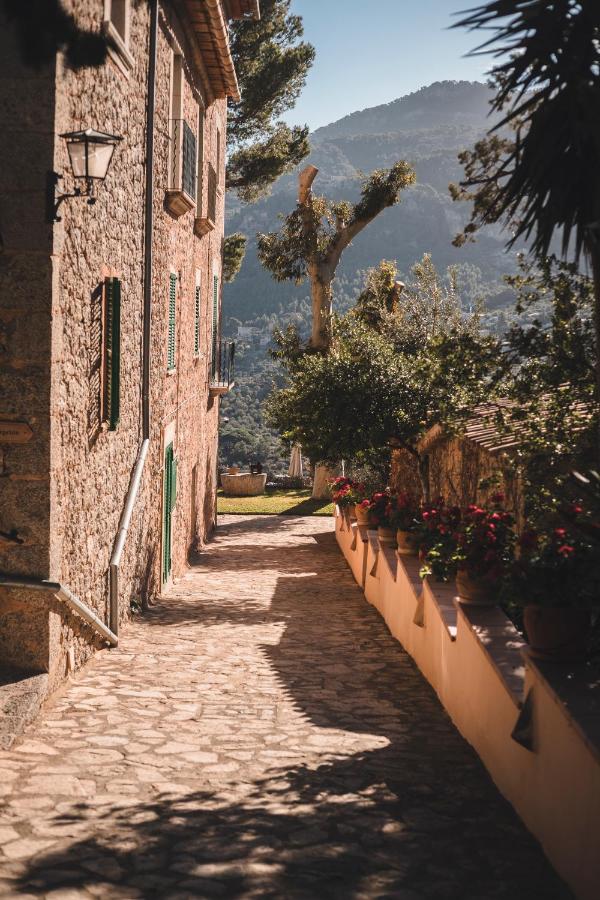 Unique Agroturismos in Mallorca 18 A narrow stone pathway beside a rustic building with green shutters, typical of agroturismos in Mallorca, is lined with potted flowers and leads toward distant mountains and trees under a clear, sunny sky.