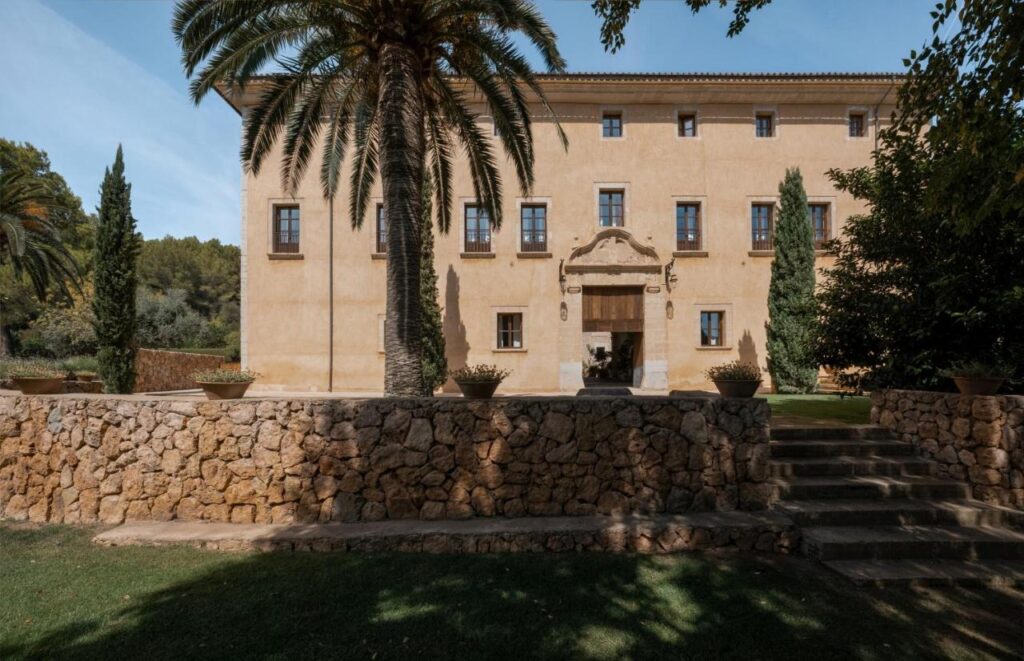 Unique Agroturismos in Mallorca 36 A large, beige villa typical of agroturismos in Mallorca, with multiple windows and a central arched entrance, is surrounded by palm trees and greenery. A stone wall and steps lead up to the entrance from a well-kept lawn.