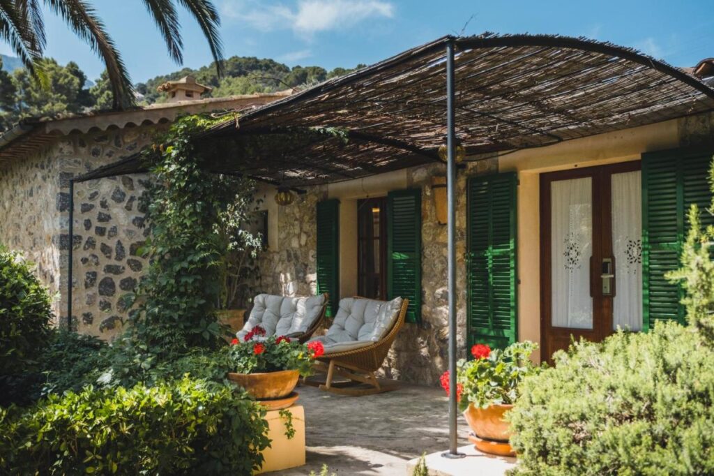 Unique Agroturismos in Mallorca 21 A stone cottage with green shutters and a vine-covered pergola shelters cushioned chairs on a sunny patio, surrounded by lush greenery and red flowers—an inviting scene typical of agroturismos in Mallorca.