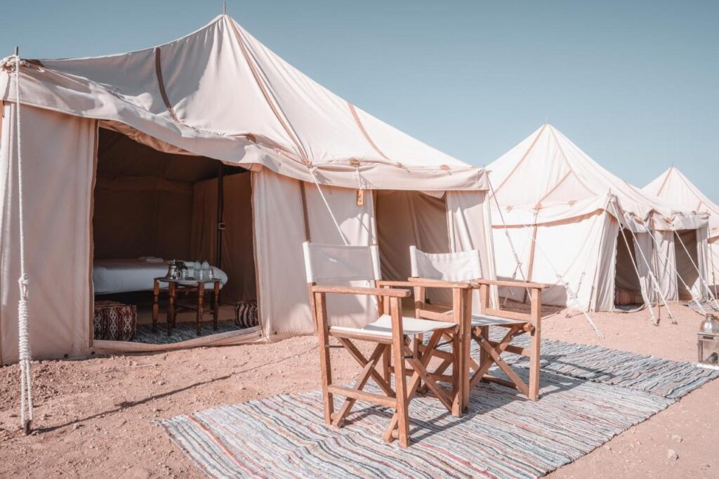 8 SPECTACULAR Agafay Desert Camps 37 Three beige canvas tents are set up on sandy ground at one of the Agafay Desert camps, with a small table and two wooden chairs arranged on a striped rug outside the front tent, which has its flap open to reveal a neatly made bed. The sky is clear and blue.