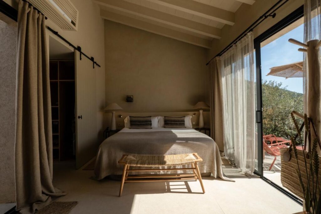 Unique Agroturismos in Mallorca 7 A cozy bedroom inspired by agroturismos in Mallorca features a large bed, neutral bedding, bedside lamps, and a wooden bench. Sunlight streams through glass doors with sheer curtains, leading to a patio with a chair and outdoor umbrella.