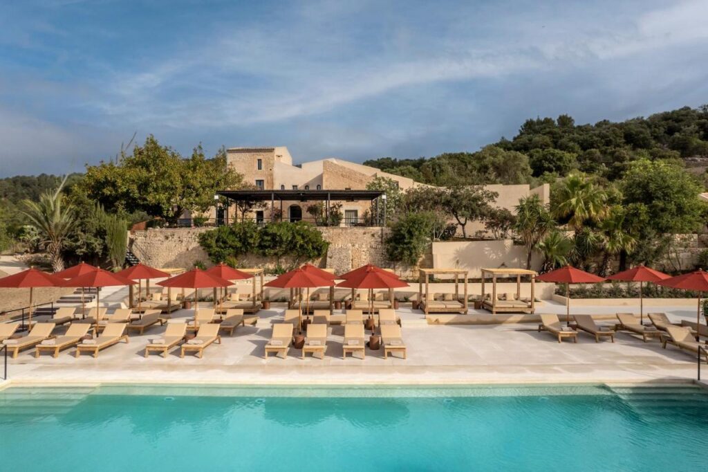 Unique Agroturismos in Mallorca 6 A luxurious outdoor pool at one of the finest agroturismos in Mallorca, with rows of beige lounge chairs and red umbrellas, set before a rustic stone building surrounded by lush greenery and trees under a partly cloudy sky.