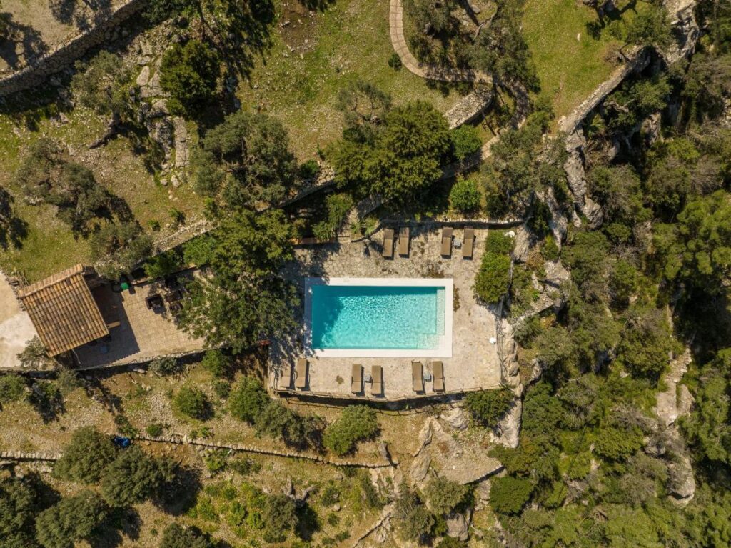 Unique Agroturismos in Mallorca 40 Aerial view of a rectangular swimming pool surrounded by lounge chairs and stone patio, nestled among trees and rocky, terraced hillsides with a small building nearby—an idyllic scene typical of agroturismos in Mallorca.