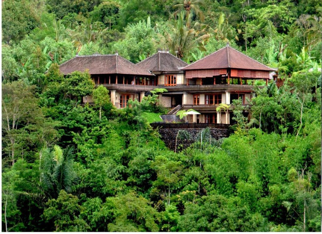 13 Most UNIQUE Places to Stay in Ubud 51 A large, multi-level villa with wooden accents and thatched roofs is nestled in lush, dense tropical greenery on a hillside—one of the unique places to stay in Ubud, surrounded by tall trees and vibrant foliage.