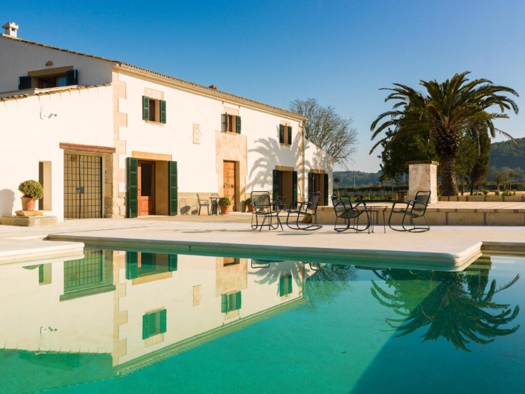 Unique Agroturismos in Mallorca 28 A large white villa with green shutters sits beside a clear swimming pool—a perfect example of agroturismos in Mallorca. Metal chairs and tables line the patio, surrounded by palm trees and distant hills under a clear blue sky.