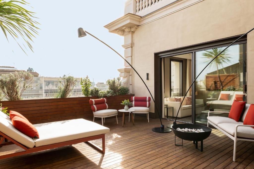 Top Barcelona Hotels with Rooftop Pool 48 A stylish outdoor terrace at Barcelona Hotels with Rooftop Pool features wooden flooring, modern lounge chairs with red and white cushions, a fire pit, a wooden bench, and potted plants, with large glass doors opening to an indoor space.