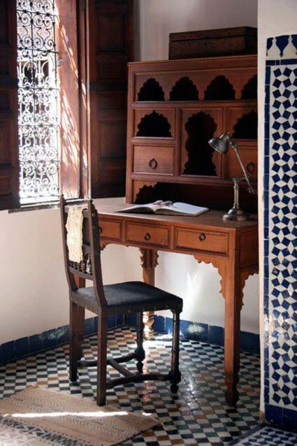 7 UNIQUE Riads in Fez You Won't Forget 6 A wooden desk and chair sit by a window with ornate ironwork in a room with patterned blue and white tile walls, echoing the timeless elegance of riads in Fez. Sunlight streams in, illuminating an open book, lamp, and shelves on the desk.
