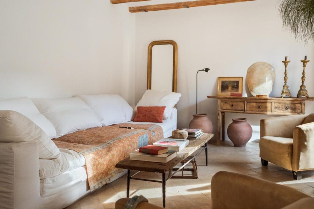 Unique Agroturismos in Mallorca 17 A cozy living room inspired by agroturismos in Mallorca features a white sofa, tan throw blanket, wooden coffee table, vintage decor, pottery, a framed mirror, and two beige armchairs. Sunlight brightens the warm, rustic space.
