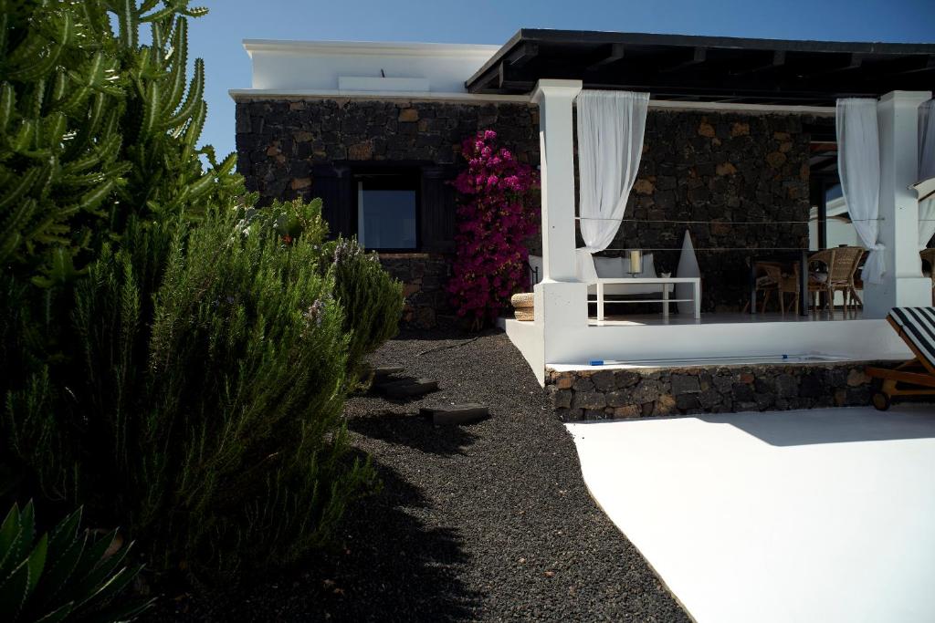 Adults Only Hotels in Fuerteventura 18 A modern house with dark stone walls, white trim, and a shaded patio with white curtains—reminiscent of the tranquil style found at adults only hotels in Fuerteventura. Pink flowers bloom near the patio, and green bushes edge the gravel path to the entrance.