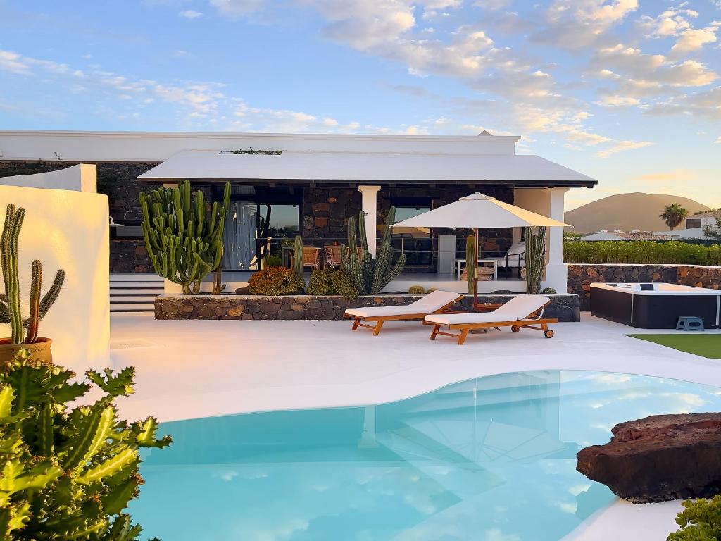 Adults Only Hotels in Fuerteventura 19 A modern villa with white walls, large windows, and stone accents, featuring a pool, sun loungers, umbrella, cacti, and a hot tub in a landscaped outdoor area at sunset—perfect for those seeking adults only hotels in Fuerteventura.