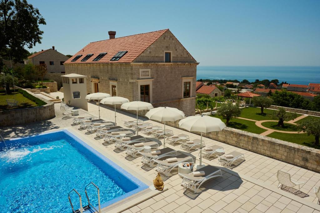 Most Unique Boutique Hotels in Dubrovnik 21 A stone villa with a red-tiled roof overlooks a blue swimming pool lined with white lounge chairs and umbrellas, set in a landscaped yard with gardens—exuding the charm of boutique hotels in Dubrovnik, with sea views in the distance.