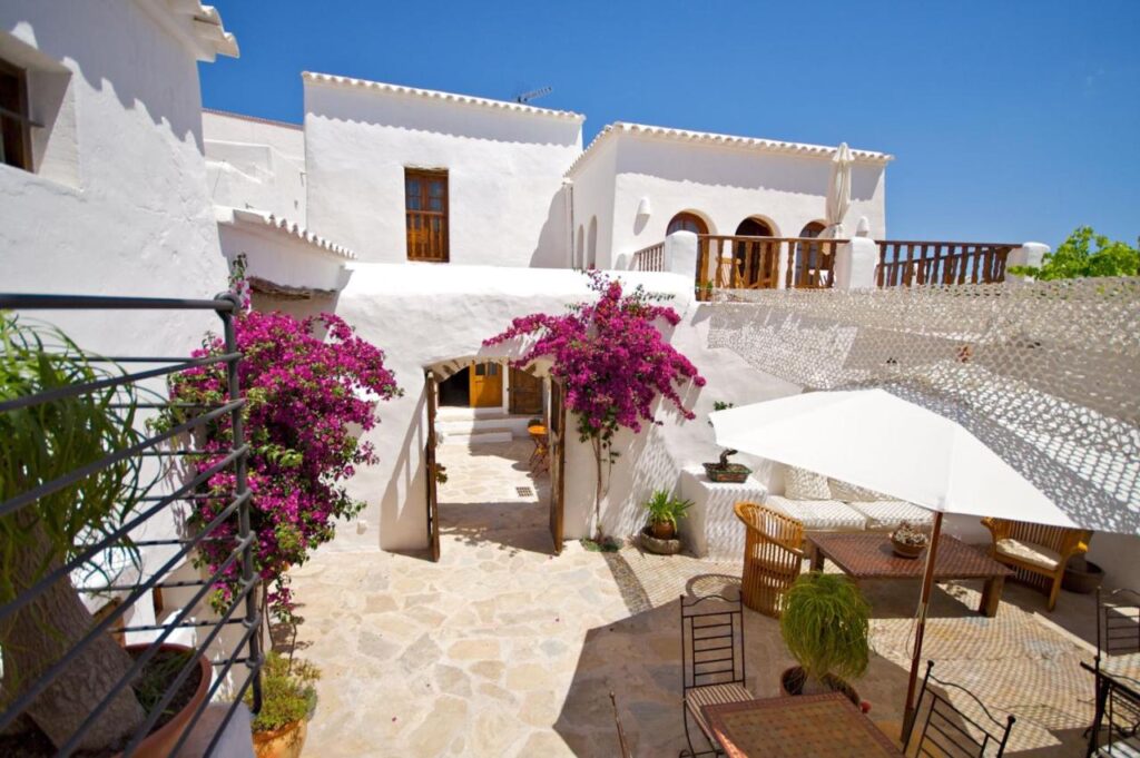 10 AUTHENTIC Finca Hotels in Ibiza 30 Sunlit courtyard of a Mediterranean-style white stucco house, resembling the charm of finca hotels in Ibiza, with flowering bougainvillea, potted plants, an outdoor dining table, chairs, and a large white umbrella under a clear blue sky.