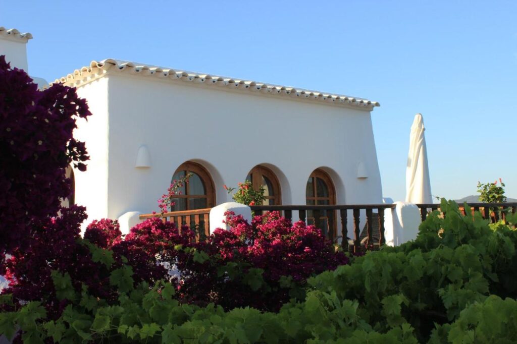 10 AUTHENTIC Finca Hotels in Ibiza 33 A white Mediterranean-style house with arched windows and a tiled roof, reminiscent of the charming finca hotels in Ibiza, is surrounded by lush greenery and blooming pink flowers under a clear blue sky.
