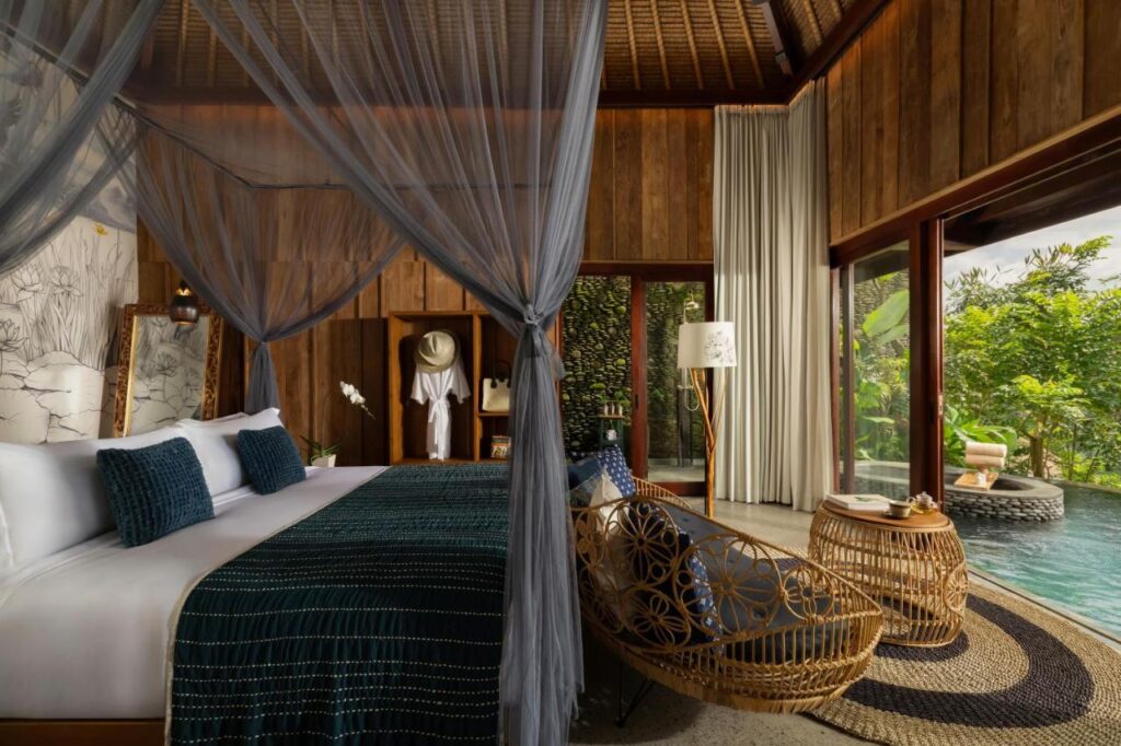 13 Most UNIQUE Places to Stay in Ubud 40 A luxurious tropical bedroom with a canopy bed, dark blue bedding, wooden walls, wicker furniture, and floor-to-ceiling windows opening to a pool and lush greenery—one of the unique places to stay in Ubud.