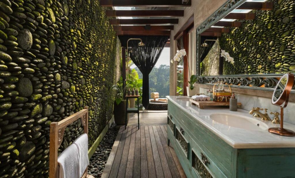 13 Most UNIQUE Places to Stay in Ubud 38 A luxurious bathroom with moss-covered stone walls, a large mirror above a turquoise vanity, wooden floors, and open views to lush greenery—one of the unique places to stay in Ubud. Natural light fills the serene space.
