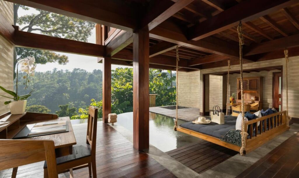 13 Most UNIQUE Places to Stay in Ubud 41 Open, airy room with wooden floors and beams, featuring a hanging daybed over a reflective pool—a perfect example of unique places to stay in Ubud. Enjoy a desk with an orchid and lush greenery through large open windows.
