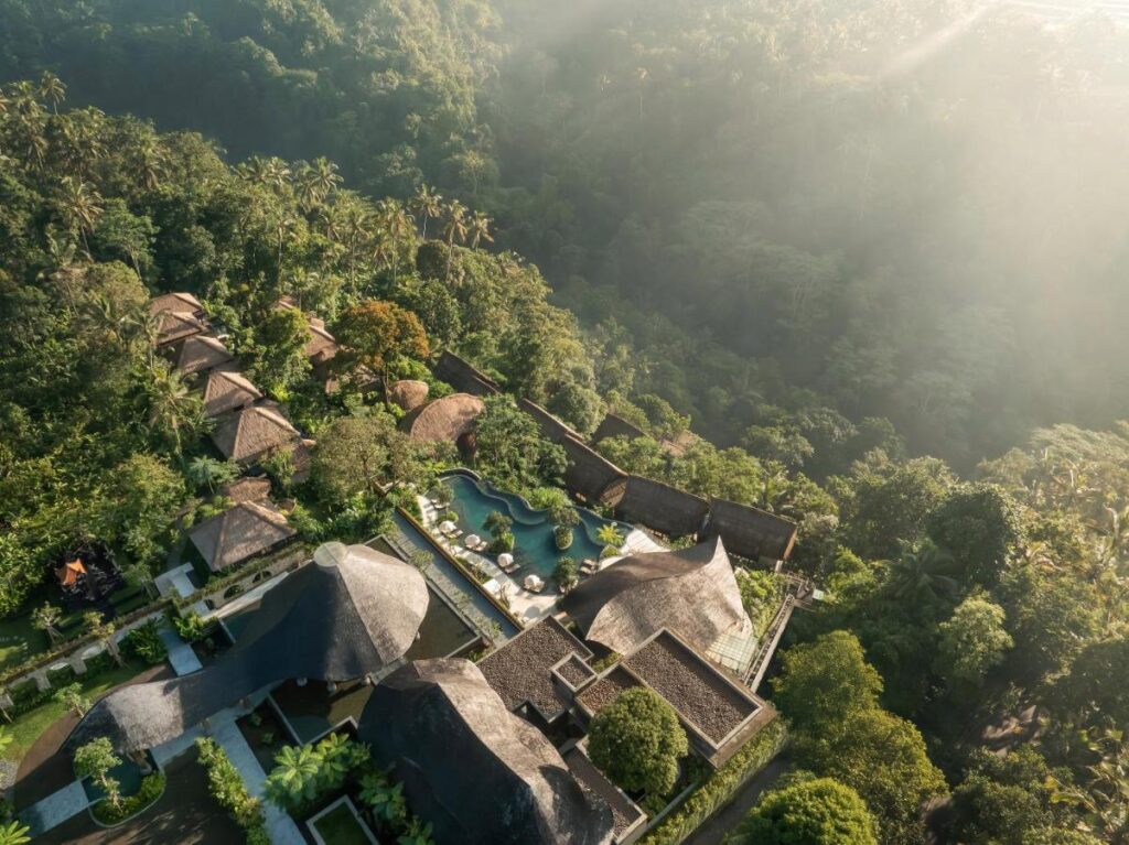13 Most UNIQUE Places to Stay in Ubud 39 Aerial view of a luxury resort with thatched-roof villas, a curving pool, and lush tropical forest surrounding the property under soft sunlight—one of the most unique places to stay in Ubud.