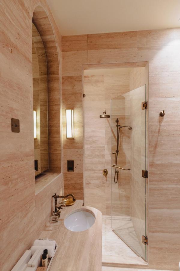 9 Best Hotels in Saudi Arabia: Unique Stays Across the Country 5 A modern bathroom in one of the Best Hotels in Saudi Arabia features beige marble walls, a built-in sink, a large mirror, glass shower enclosure, and elegant brass fixtures. A wall light beside the mirror highlights neatly arranged toiletries.