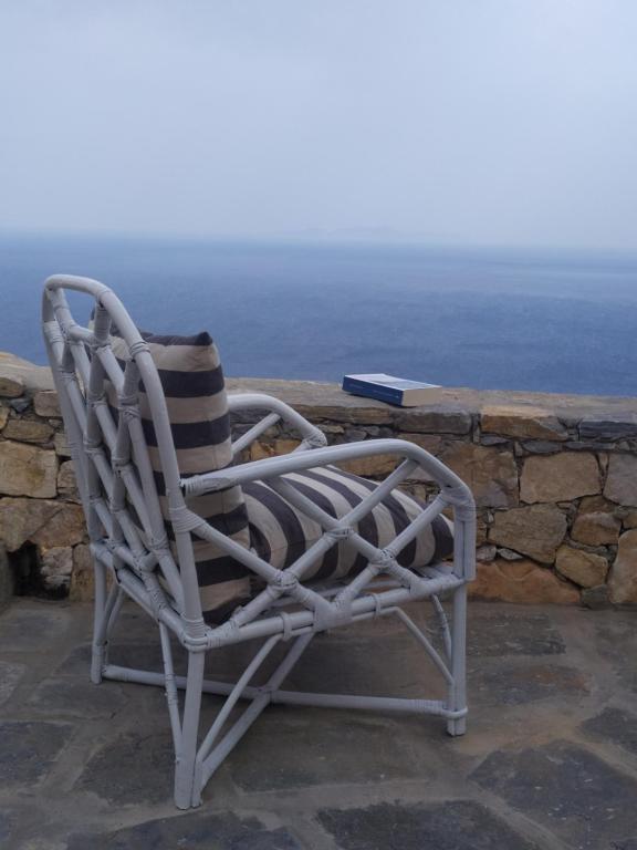 Authentic Hotels in Sikinos, Cyclades 5 A wicker chair with striped cushions sits on a stone patio overlooking a calm, blue sea—reminiscent of the tranquil charm found at hotels in Sikinos. A closed book rests on the low stone wall in the background beneath a cloudy sky.